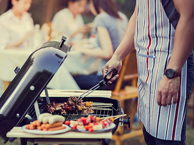 Man grilling food outside with his family sitting at a table in the background.