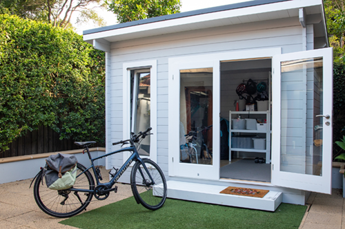 An open double-door modern shed in a backyard, with storage shelves visible inside. A bicycle is parked in front of the shed on artificial green turf, surrounded by wooden fencing and greenery.