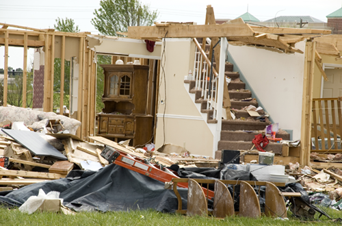 Image of a partially demolished house with visible wooden frames and scattered debris. An intact staircase leads to the second floor, and amidst the wreckage, a piece of wooden furniture stands undamaged. The scene is set against an overcast sky, indicating a residential area possibly affected by severe weather.