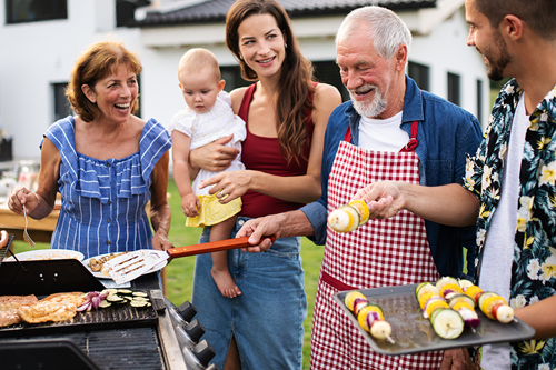 Man grilling food outside with his family sitting at a table in the background.